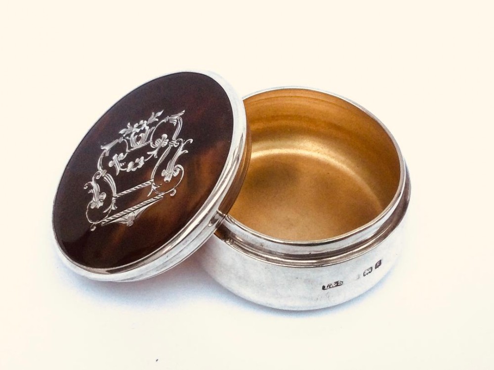 A circular Edwardian sterling silver pill box displayed open alongside a silver-mounted lid featuring a dark brown faux tortoiseshell inset with silver cartouche inlay.