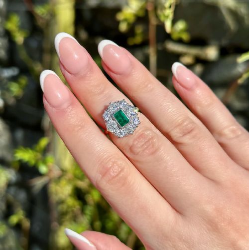 fig 5. An antique Edwardian cluster ring featuring a central rectangular step-cut green emerald framed by eight old-cut diamonds in a white metal setting.