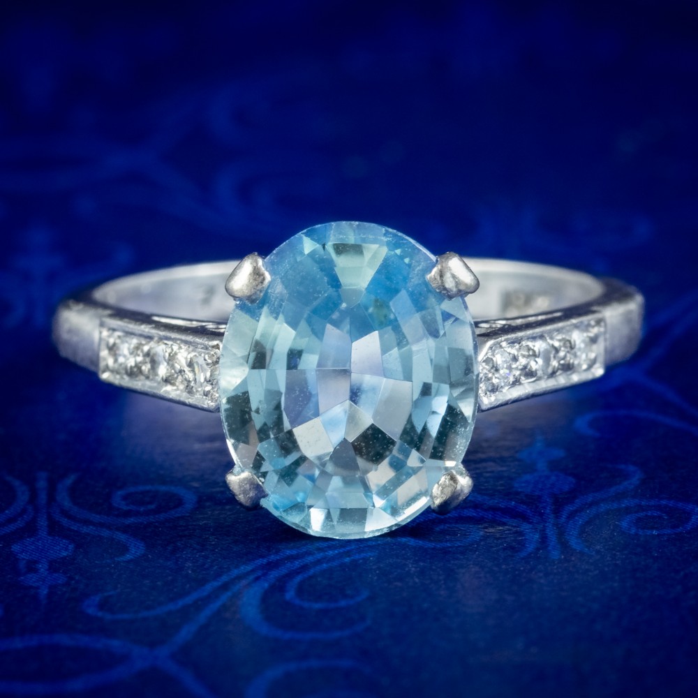 An oval-cut light blue aquamarine gemstone set in a four-claw mount with channel-set diamond shoulders on a silver-coloured metal band.