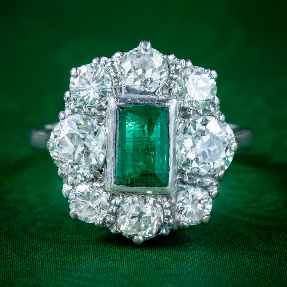 An antique Edwardian cluster ring featuring a central rectangular step-cut green emerald framed by eight old-cut diamonds in a white metal setting.