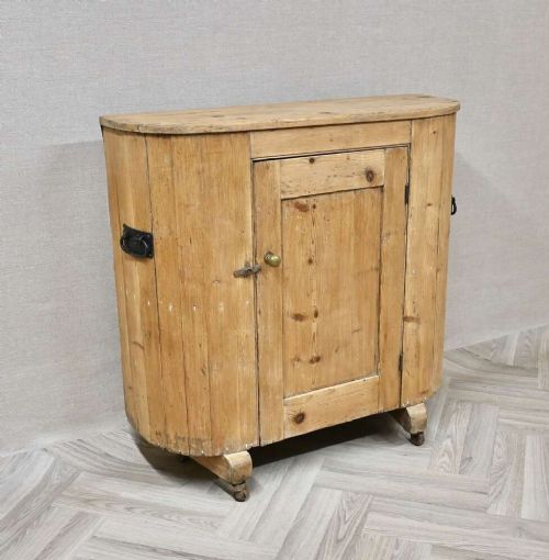 An antique natural pine huffer cupboard featuring a curved front, single panelled door with a brass knob, iron latch, side handles, and short wooden legs on metal castors. View II