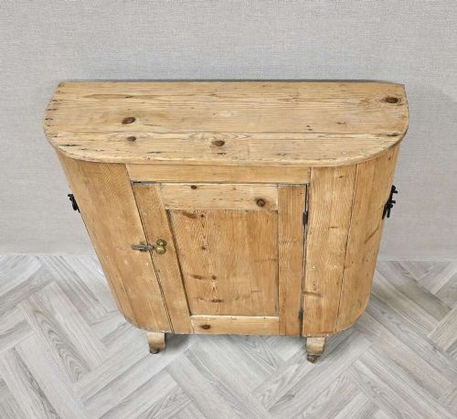 An antique natural pine huffer cupboard featuring a curved front, single panelled door with a brass knob, iron latch, side handles, and short wooden legs on metal castors. View III