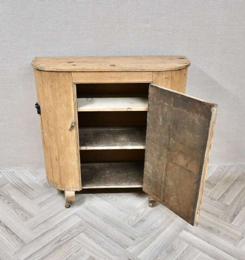 An antique natural pine huffer cupboard featuring a curved front, single panelled door with a brass knob, iron latch, side handles, and short wooden legs on metal castors. View IV