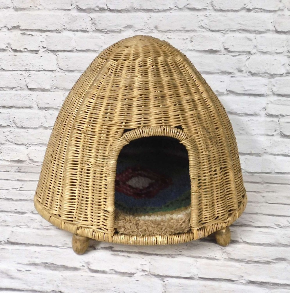 A dome-shaped woven wicker pet basket with an arched entrance and a cushioned interior rests against a white brick background.
