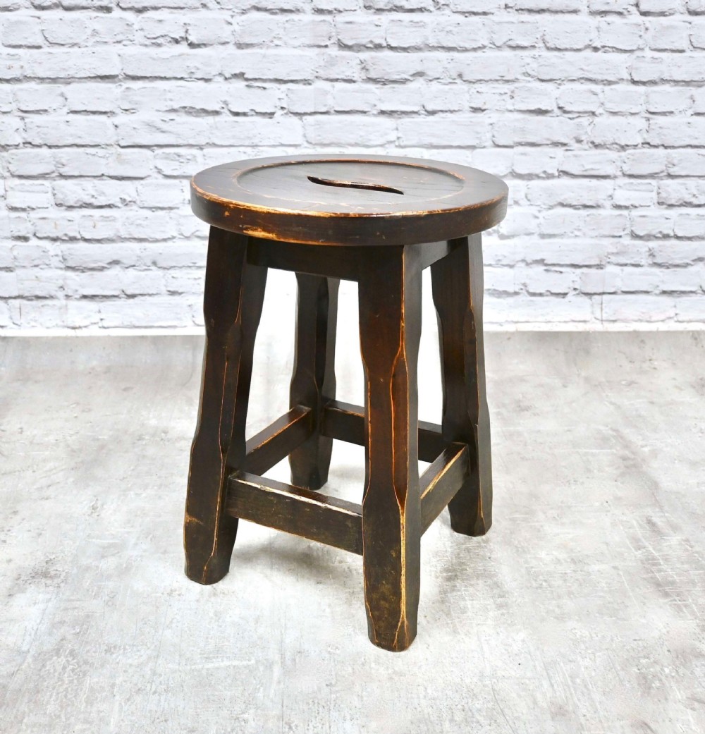 A dark brown Arts and Crafts solid oak stool with a round seat featuring a central hand slot and faceted tapered legs. View VII