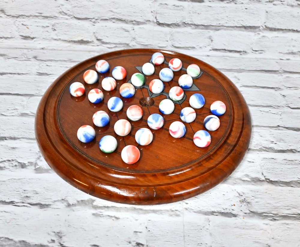 A circular nineteenth-century wooden solitaire game board featuring turned edges and incised guidelines, set with multicoloured swirl glass marbles.