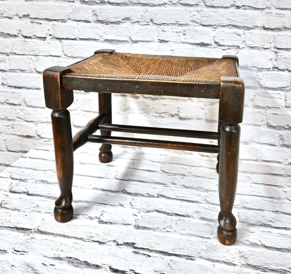 A dark brown wooden Arts and Crafts footstool featuring turned legs, horizontal stretchers, and a woven natural rush seat.