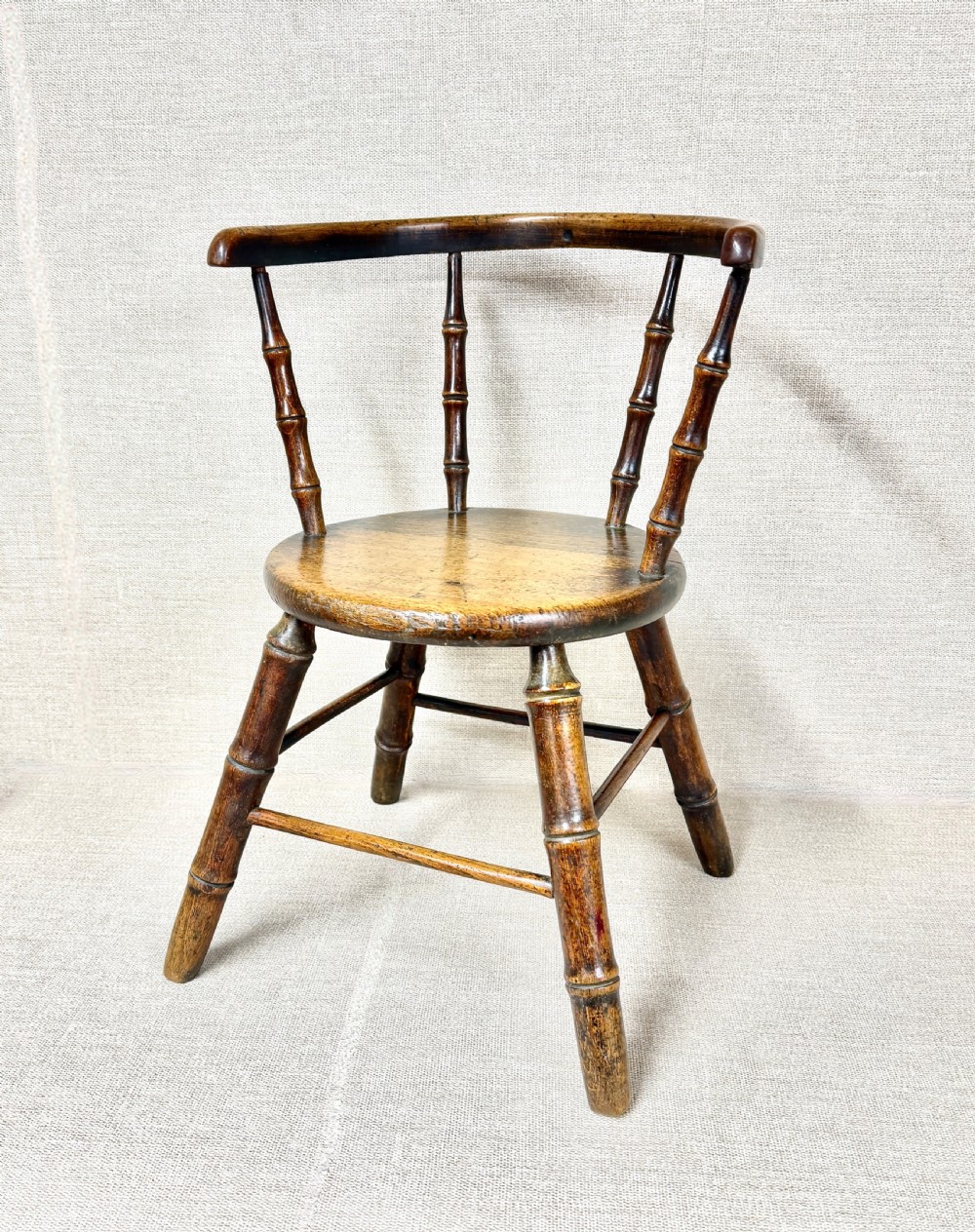 An antique dark wood childs Windsor chair featuring turned faux-bamboo spindles, a curved top rail, a solid wooden seat, and splayed turned legs with stretchers.