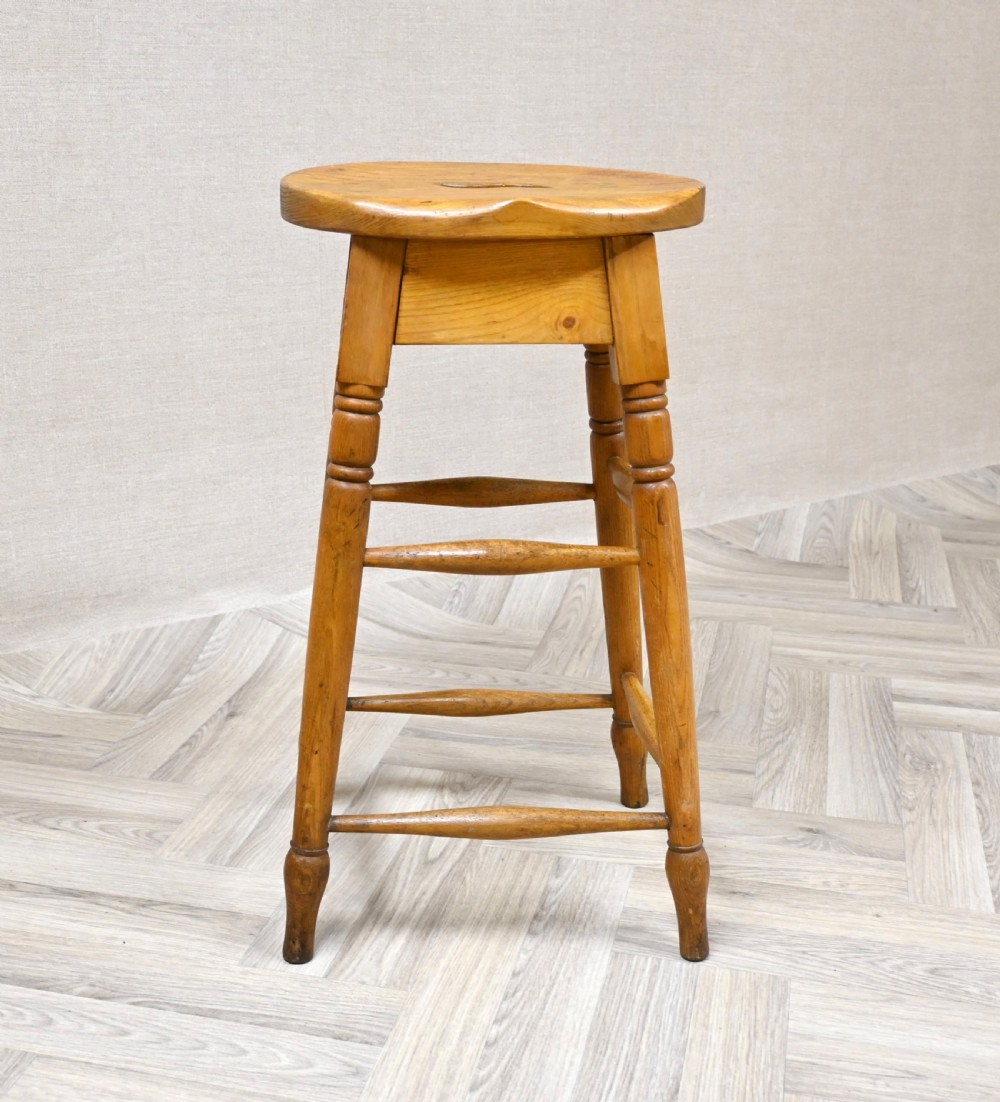 A tall 19th-century wooden bar stool featuring turned legs, lower footrest stretchers, and a rounded seat with a carved handhold slot. View V