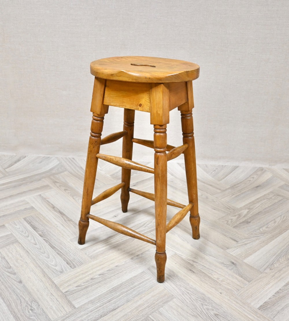 A tall 19th-century wooden bar stool featuring turned legs, lower footrest stretchers, and a rounded seat with a carved handhold slot. View VI