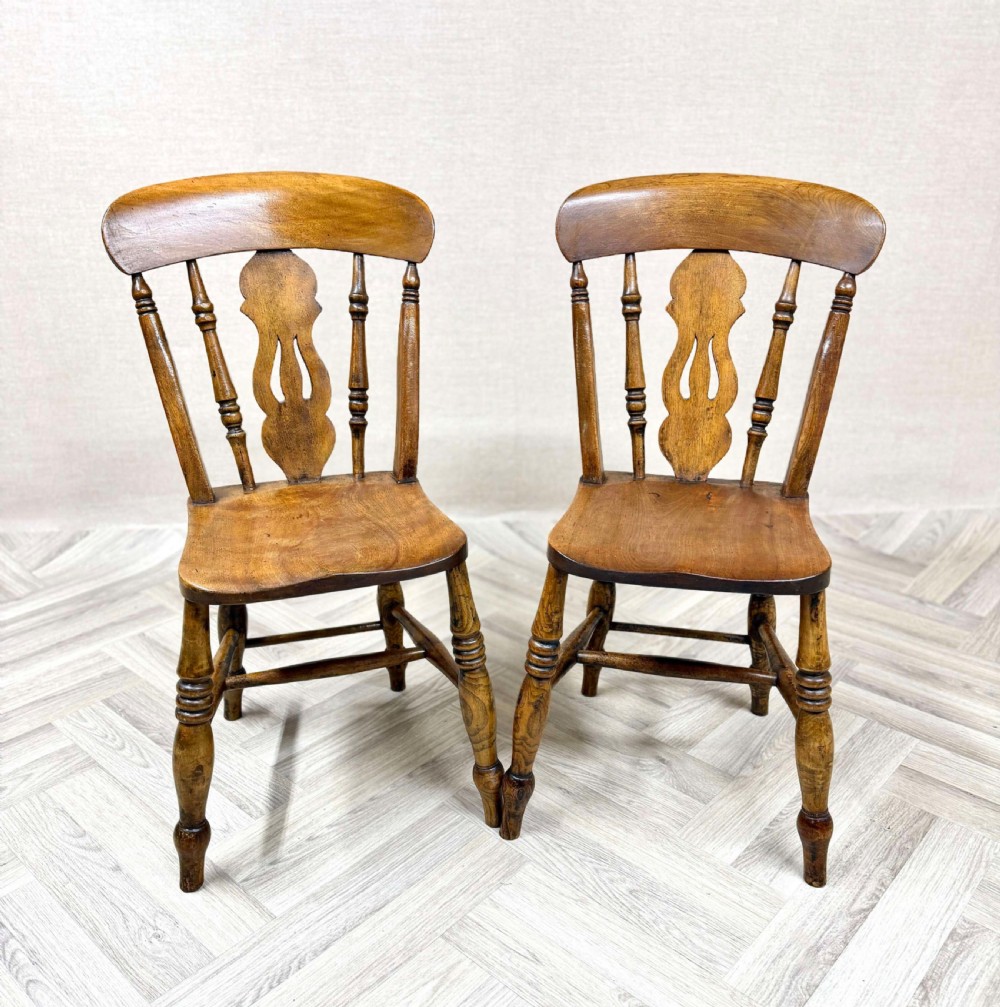 A pair of antique wooden Windsor kitchen chairs featuring carved lyreback splats, turned spindles, shaped wooden seats, and turned legs joined by stretchers.