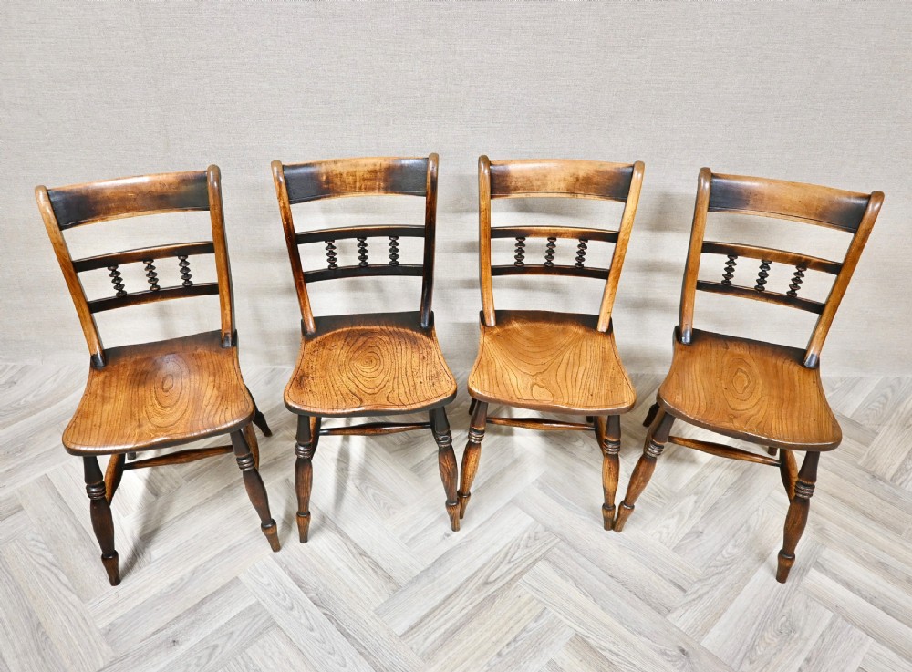A set of four 19th-century wooden Windsor kitchen dining chairs featuring shaped elm seats, bar backs with spindle details, turned legs, and H-stretchers. View IV