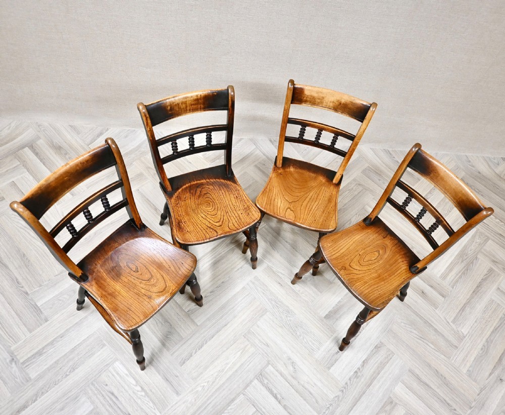 A set of four 19th-century wooden Windsor kitchen dining chairs featuring shaped elm seats, bar backs with spindle details, turned legs, and H-stretchers. View V