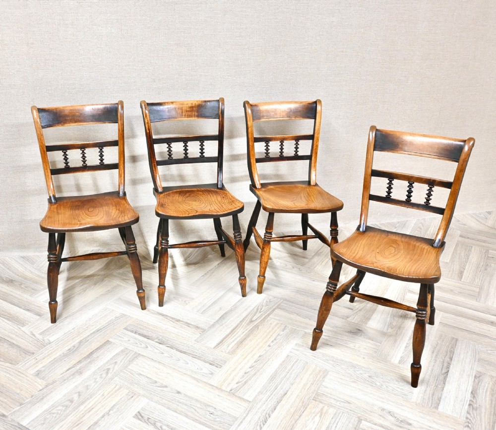 A set of four 19th-century wooden Windsor kitchen dining chairs featuring shaped elm seats, bar backs with spindle details, turned legs, and H-stretchers. View VI