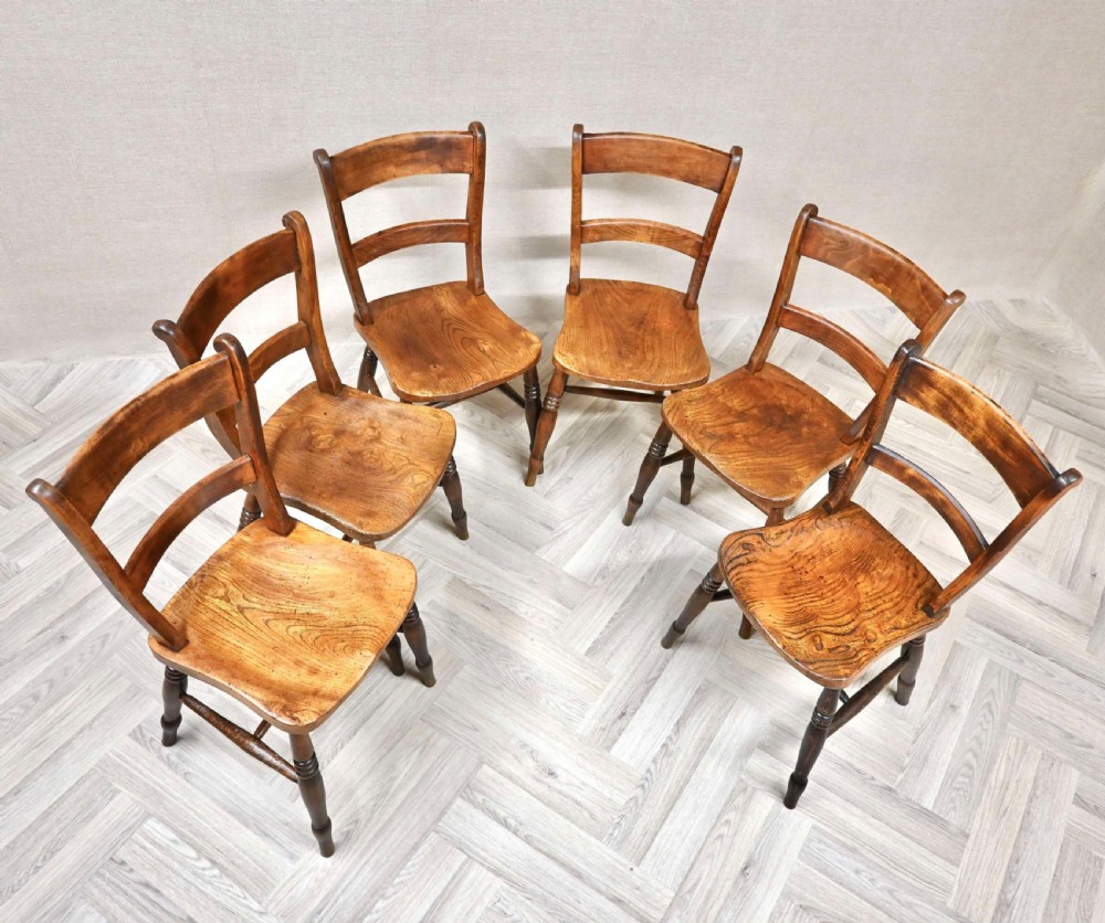 A set of six 19th-century wooden farmhouse kitchen chairs with curved horizontal back rails, figured wooden seats, and turned legs arranged in a semi-circle.