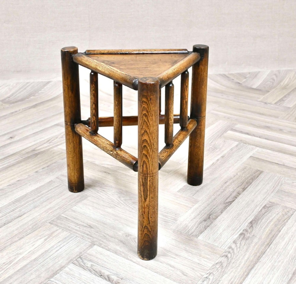 A triangular dark oak Arts and Crafts stool with three thick turned upright posts, vertical spindles, and triangular framing. View VI