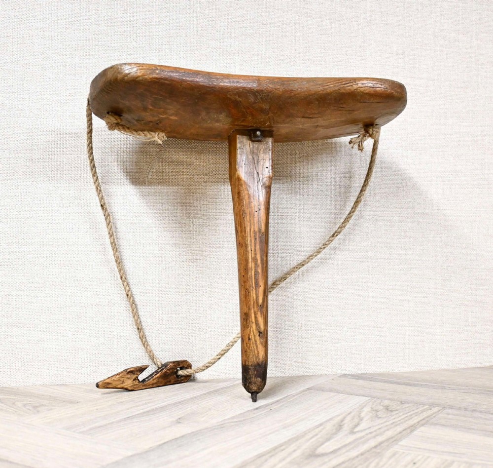 A hand-carved rustic wooden one-legged milking stool with a thick contoured seat, central tapered leg, and original hemp rope strap.