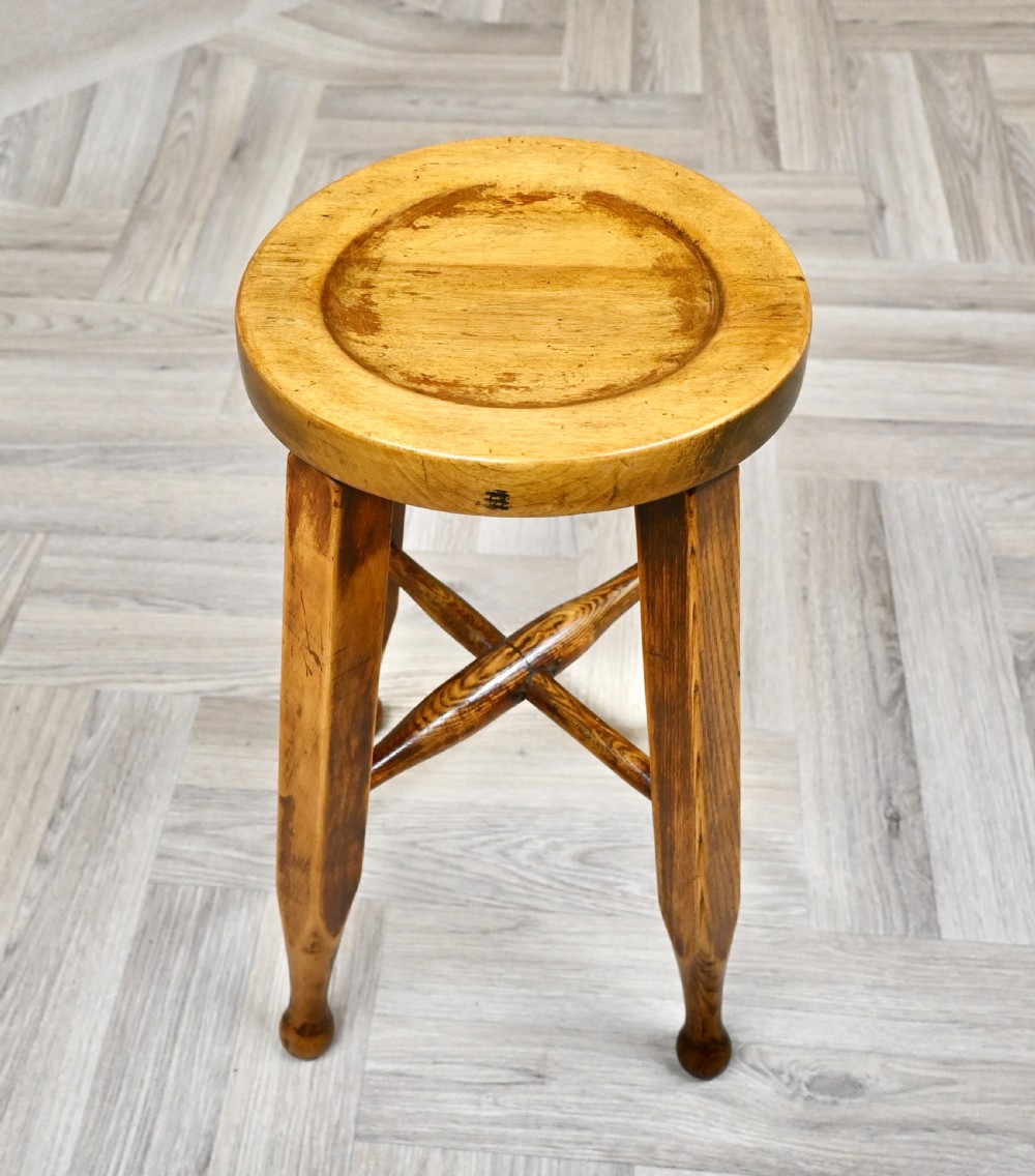 A solid wooden stool featuring a round dished seat, four angled legs with subtle turned feet, and a central cross stretcher in a warm amber finish. View II