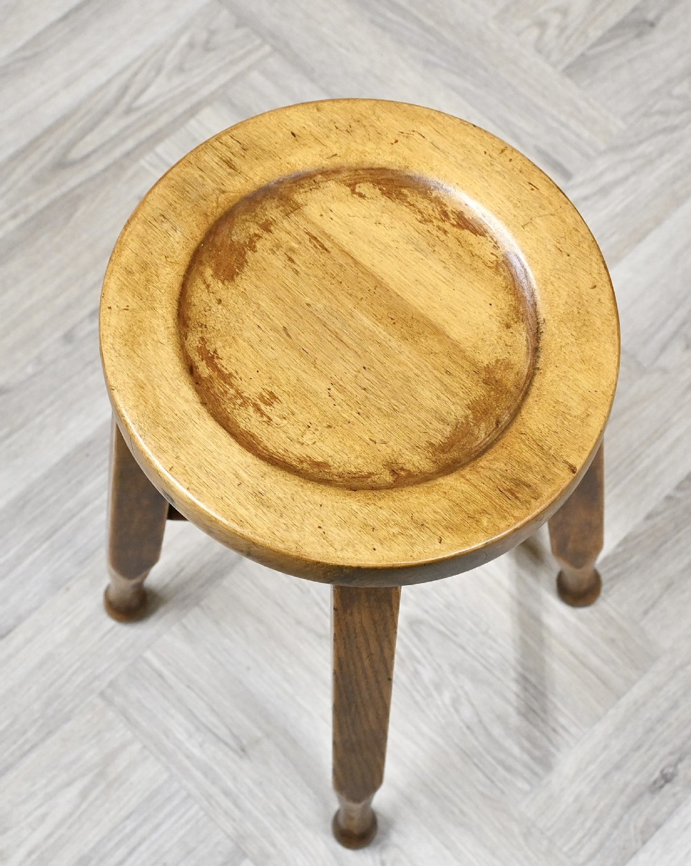 A solid wooden stool featuring a round dished seat, four angled legs with subtle turned feet, and a central cross stretcher in a warm amber finish. View III