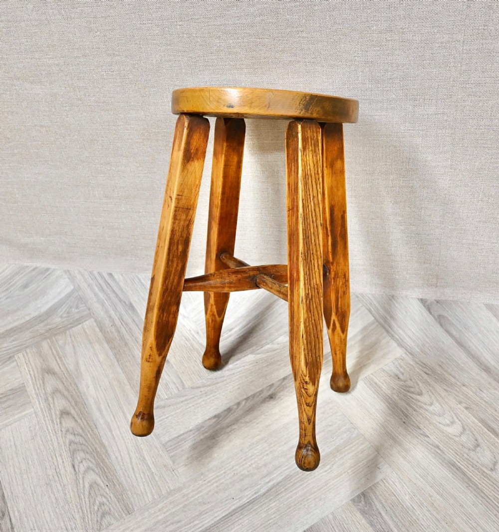 A solid wooden stool featuring a round dished seat, four angled legs with subtle turned feet, and a central cross stretcher in a warm amber finish. View IV