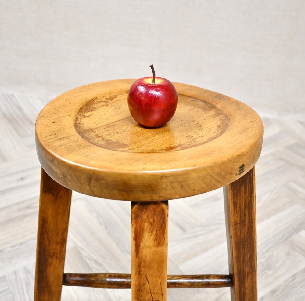 A solid wooden stool featuring a round dished seat, four angled legs with subtle turned feet, and a central cross stretcher in a warm amber finish. View V