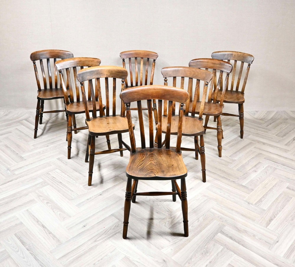 A set of eight warm brown wooden Windsor dining chairs with turned legs and curved spindle backrests arranged on a neutral herringbone floor.