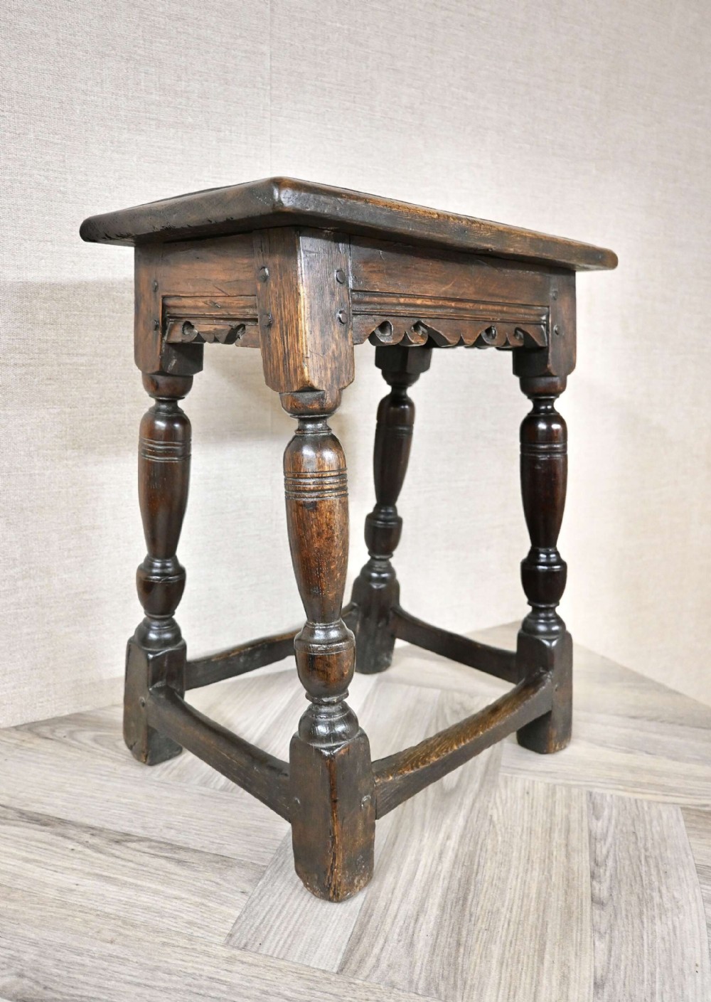 A dark oak joint stool featuring turned baluster legs, a carved apron frieze, wooden peg joinery, and low perimeter stretchers.