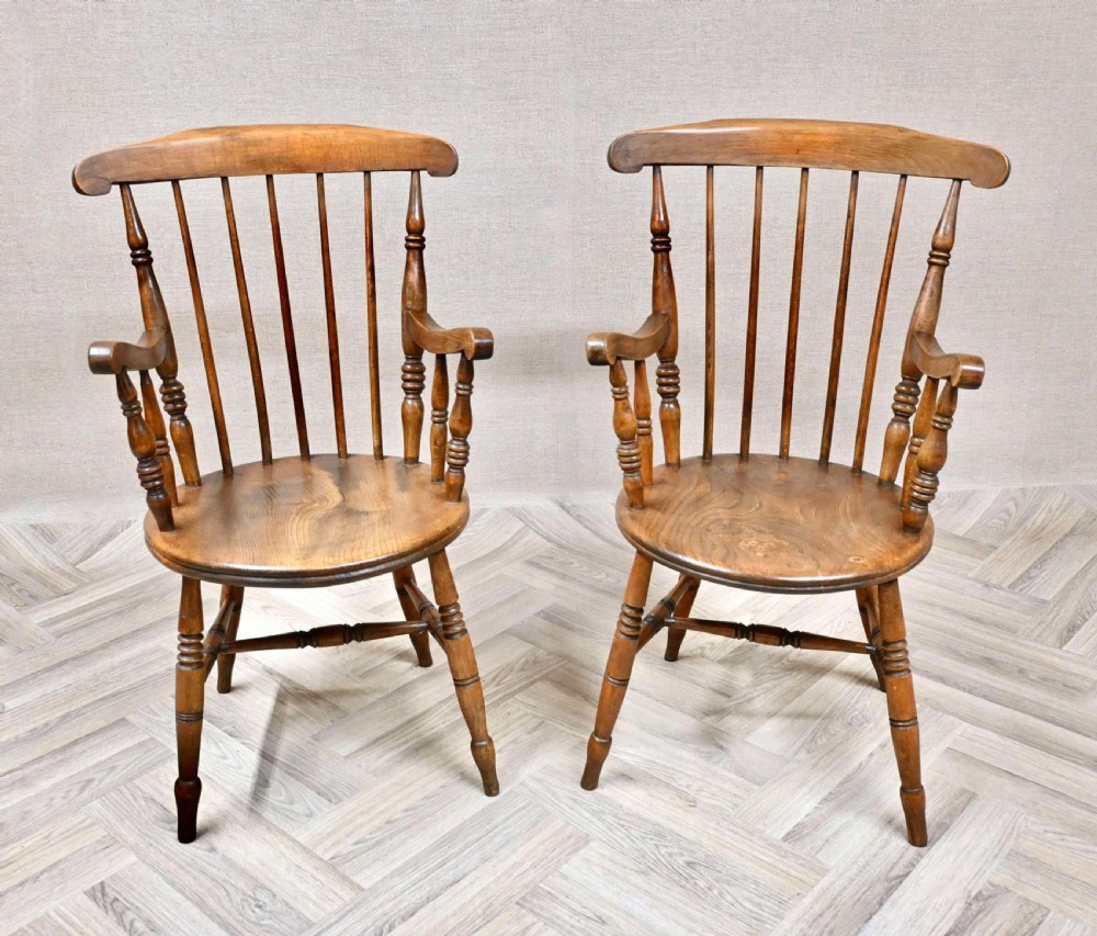 A pair of warm brown wooden Windsor carver armchairs featuring circular penny seats, turned legs, and spindle backs.