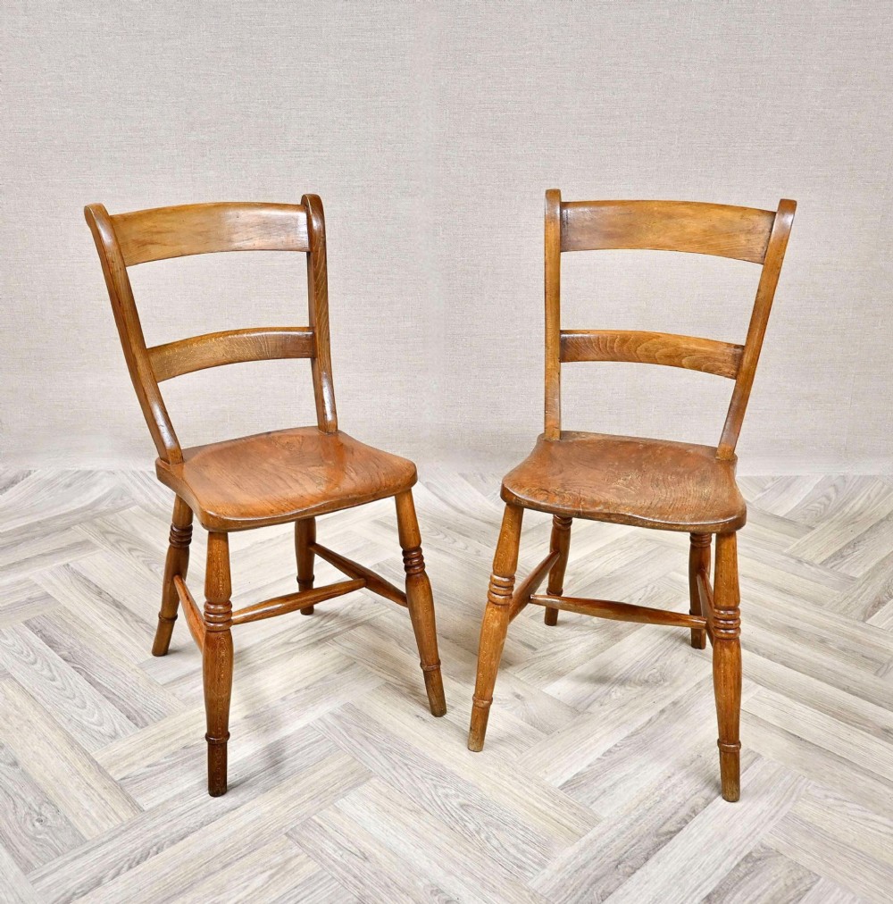A pair of antique warm brown wooden barback kitchen chairs featuring solid contoured seats, double horizontal back rails, and turned legs.
