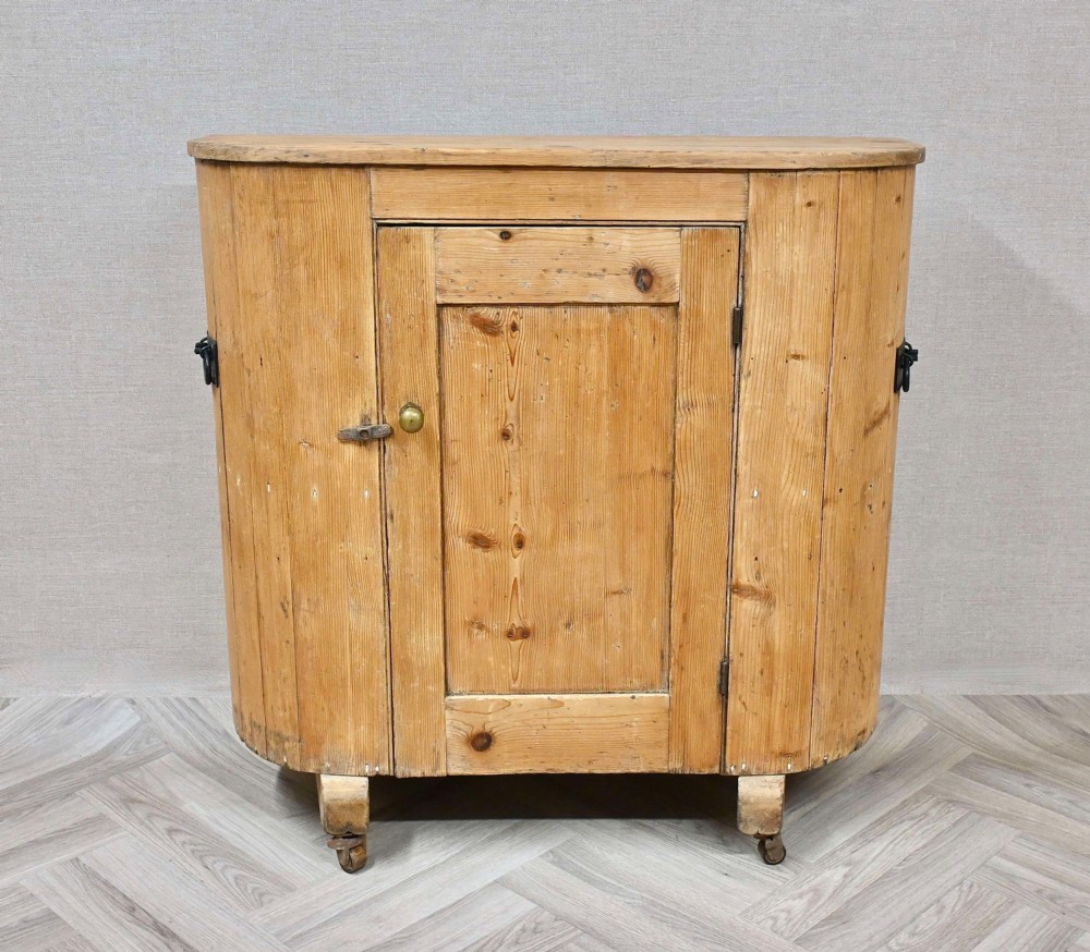 An antique natural pine huffer cupboard featuring a curved front, single panelled door with a brass knob, iron latch, side handles, and short wooden legs on metal castors.