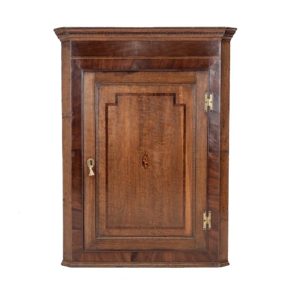 A late 18th-century Georgian oak wall-hanging corner cupboard featuring mahogany crossbanding, an inlaid central lozenge motif, a moulded top cornice, and brass H-hinges.