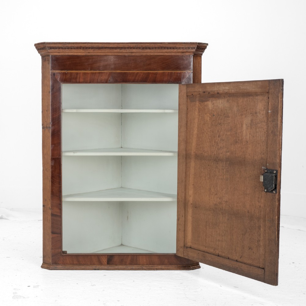 A late 18th-century Georgian oak wall-hanging corner cupboard featuring mahogany crossbanding, an inlaid central lozenge motif, a moulded top cornice, and brass H-hinges. View V