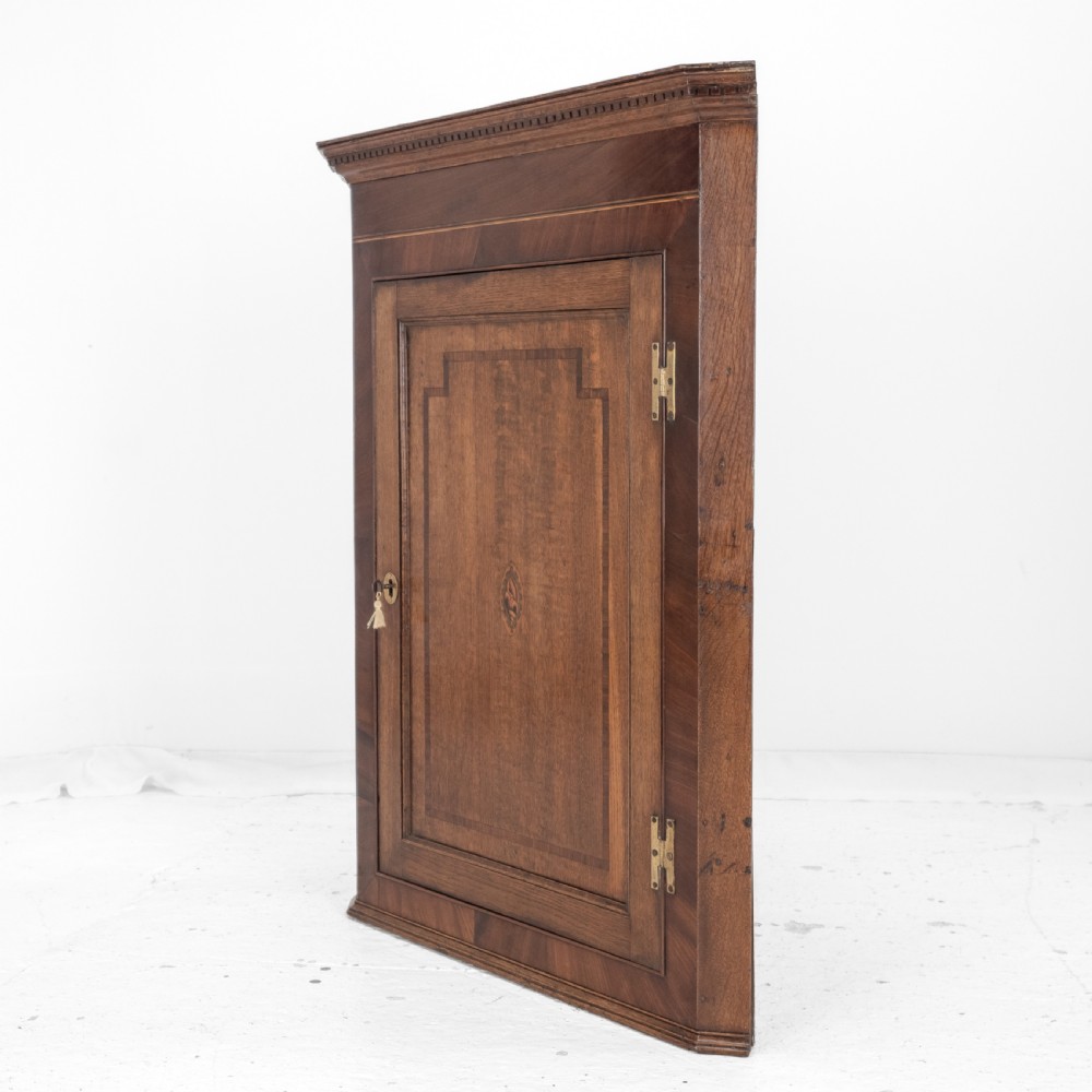 A late 18th-century Georgian oak wall-hanging corner cupboard featuring mahogany crossbanding, an inlaid central lozenge motif, a moulded top cornice, and brass H-hinges. View XV
