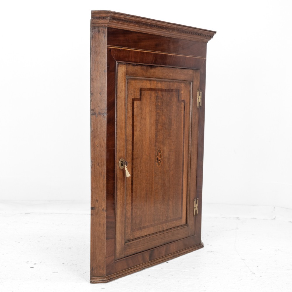 A late 18th-century Georgian oak wall-hanging corner cupboard featuring mahogany crossbanding, an inlaid central lozenge motif, a moulded top cornice, and brass H-hinges. View XVI