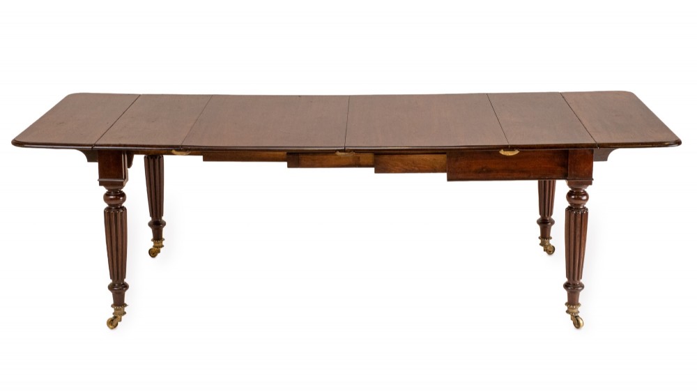An extended Georgian mahogany dining table featuring a smooth polished surface, rectangular top with rounded corners, and reeded turned legs with brass castors. View IV