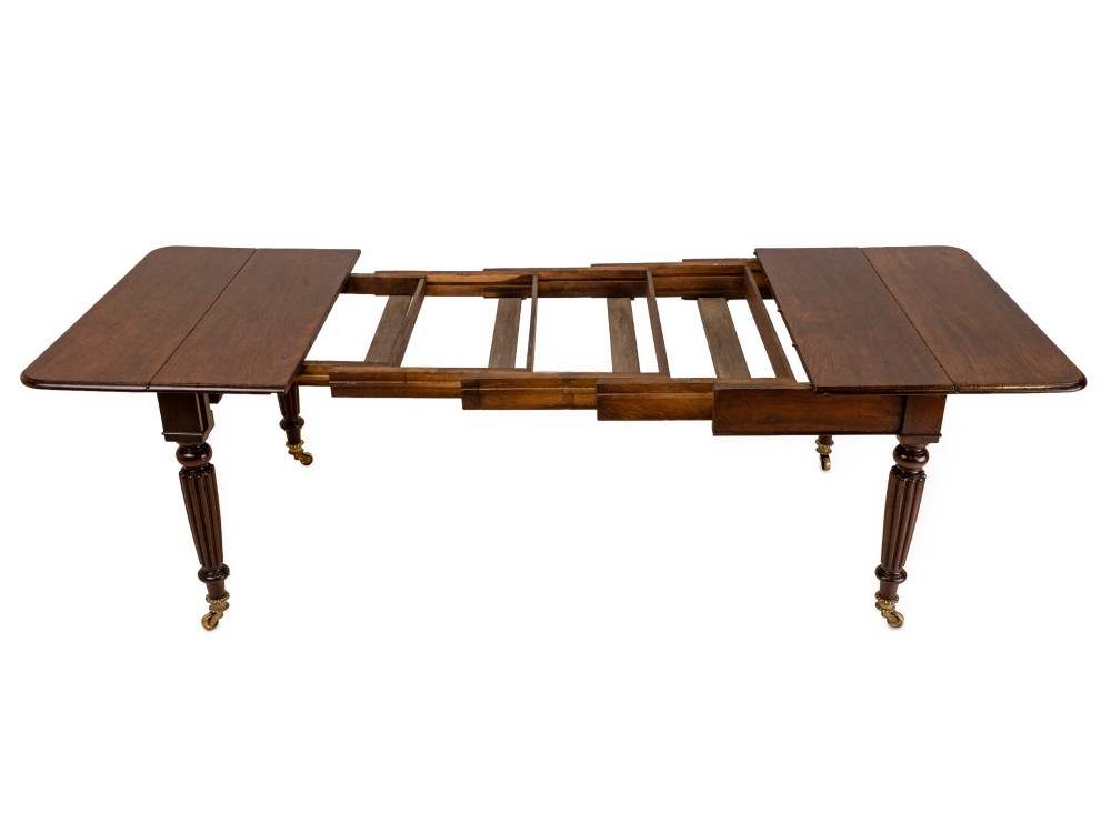 An extended Georgian mahogany dining table featuring a smooth polished surface, rectangular top with rounded corners, and reeded turned legs with brass castors. View V