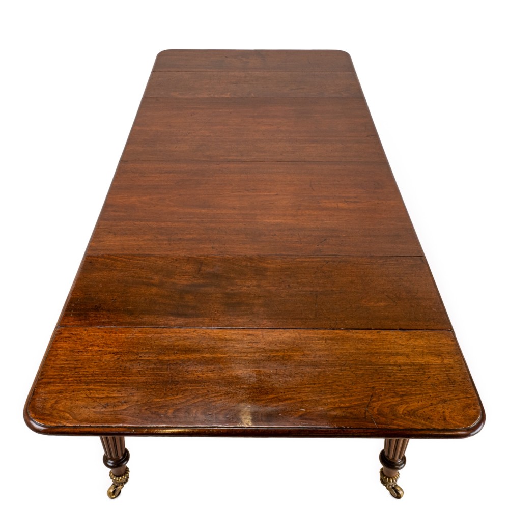 An extended Georgian mahogany dining table featuring a smooth polished surface, rectangular top with rounded corners, and reeded turned legs with brass castors. View VI