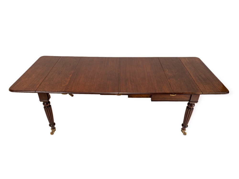 An extended Georgian mahogany dining table featuring a smooth polished surface, rectangular top with rounded corners, and reeded turned legs with brass castors. View XI