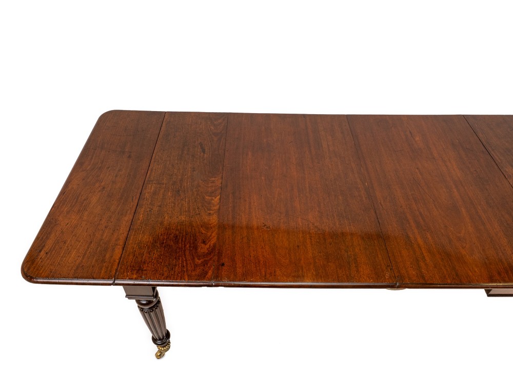 An extended Georgian mahogany dining table featuring a smooth polished surface, rectangular top with rounded corners, and reeded turned legs with brass castors. View XII