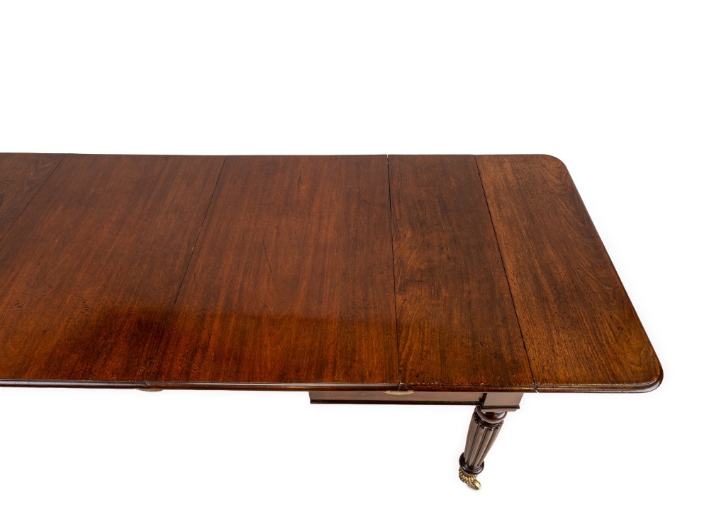 An extended Georgian mahogany dining table featuring a smooth polished surface, rectangular top with rounded corners, and reeded turned legs with brass castors. View XIII