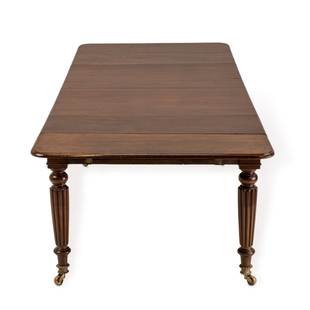 An extended Georgian mahogany dining table featuring a smooth polished surface, rectangular top with rounded corners, and reeded turned legs with brass castors. View XV