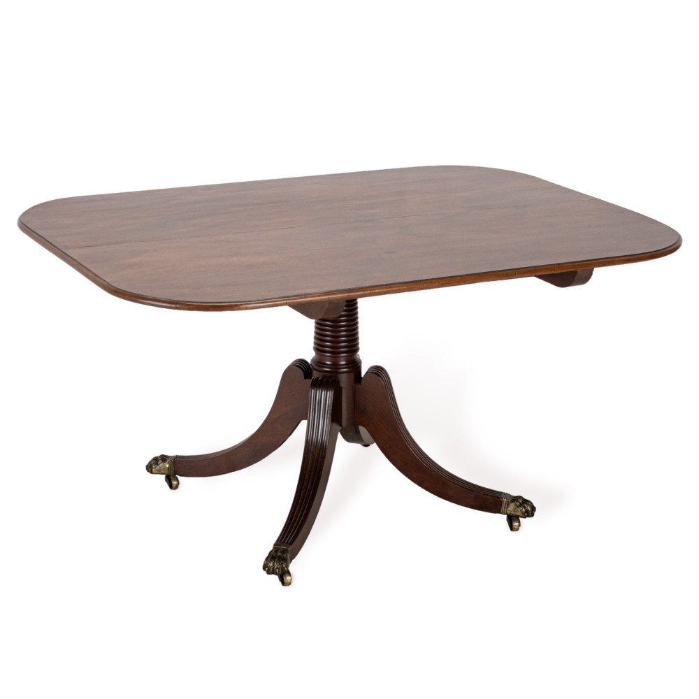 A George III mahogany dining table featuring a rectangular tilt-top surface with rounded corners set on a reeded central pedestal and four splayed legs with brass paw castors.