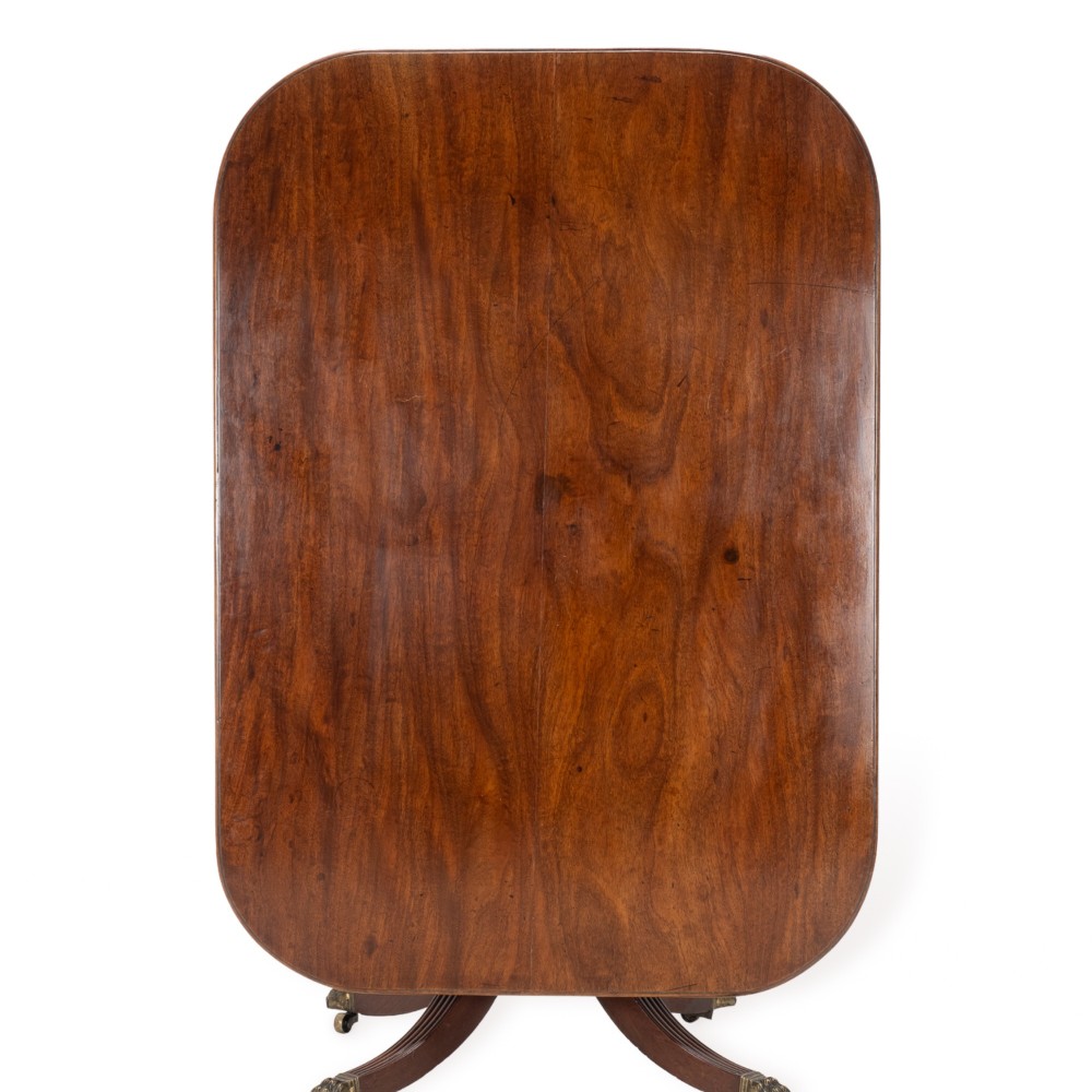 A George III mahogany dining table featuring a rectangular tilt-top surface with rounded corners set on a reeded central pedestal and four splayed legs with brass paw castors. View III