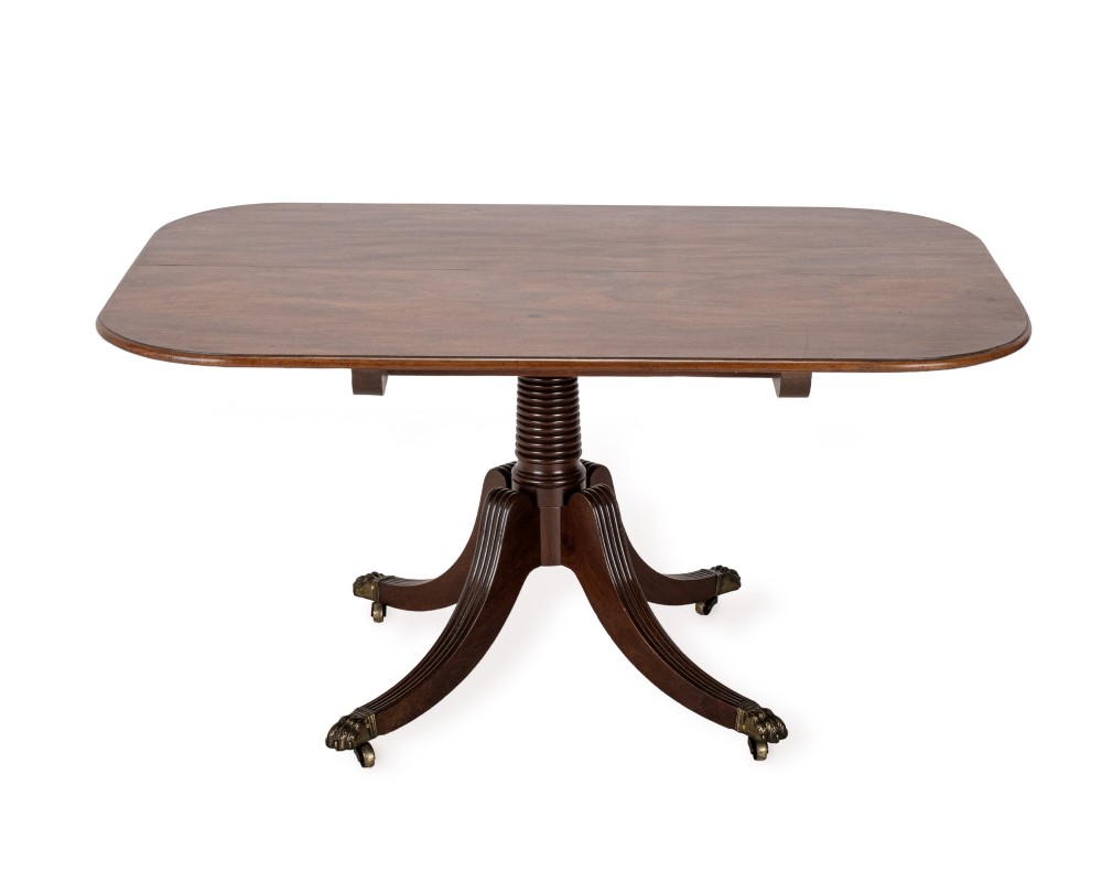 A George III mahogany dining table featuring a rectangular tilt-top surface with rounded corners set on a reeded central pedestal and four splayed legs with brass paw castors. View IV