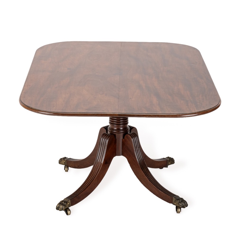 A George III mahogany dining table featuring a rectangular tilt-top surface with rounded corners set on a reeded central pedestal and four splayed legs with brass paw castors. View V
