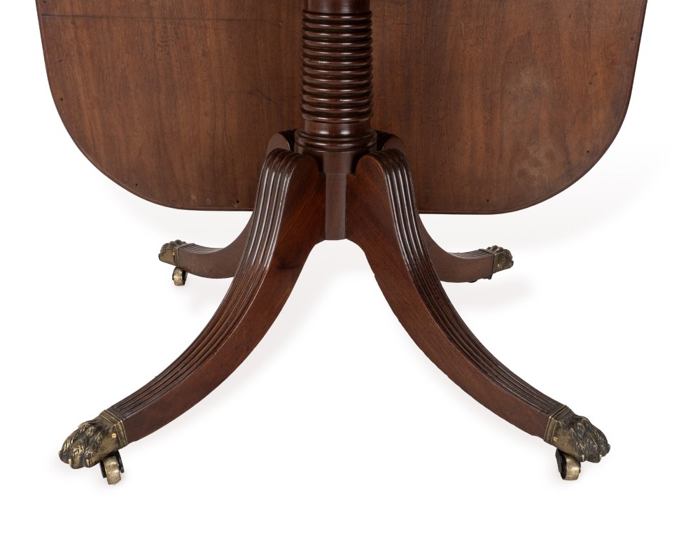 A George III mahogany dining table featuring a rectangular tilt-top surface with rounded corners set on a reeded central pedestal and four splayed legs with brass paw castors. View VI