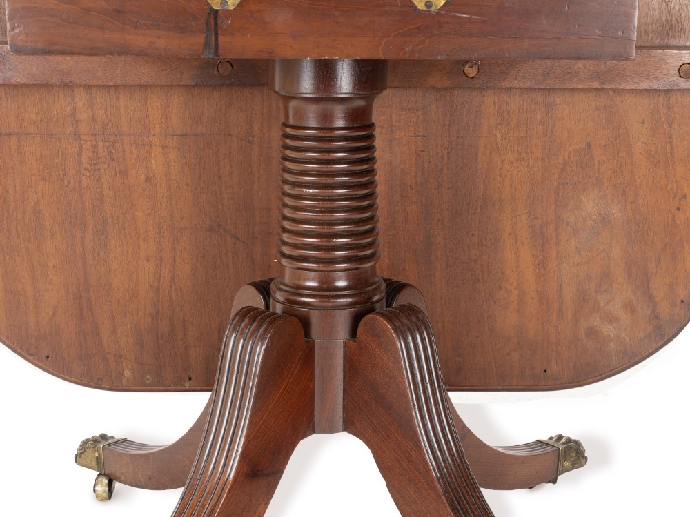 A George III mahogany dining table featuring a rectangular tilt-top surface with rounded corners set on a reeded central pedestal and four splayed legs with brass paw castors. View VII