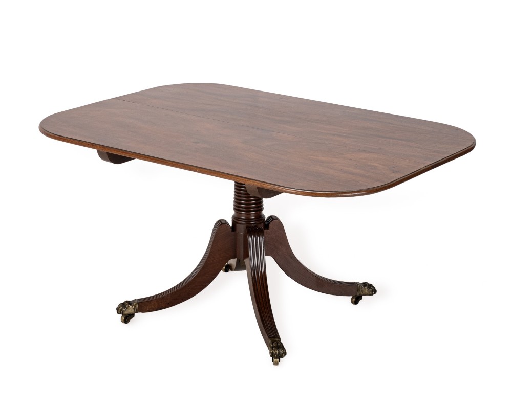 A George III mahogany dining table featuring a rectangular tilt-top surface with rounded corners set on a reeded central pedestal and four splayed legs with brass paw castors. View VIII