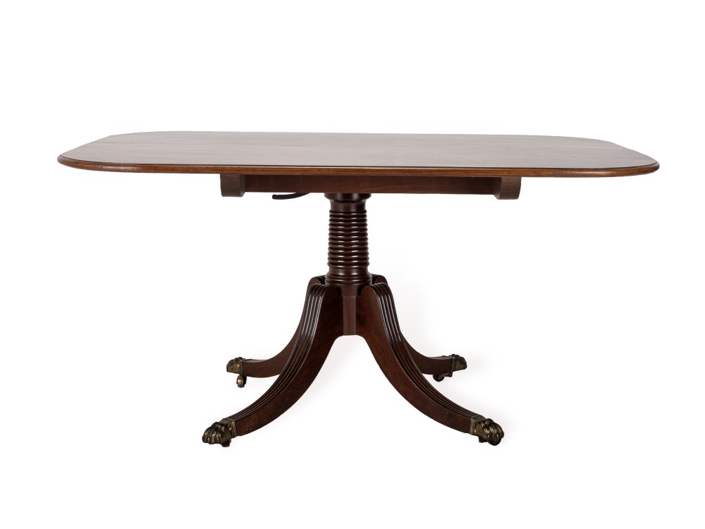 A George III mahogany dining table featuring a rectangular tilt-top surface with rounded corners set on a reeded central pedestal and four splayed legs with brass paw castors. View IX
