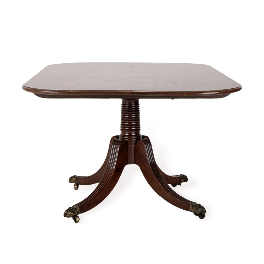 A George III mahogany dining table featuring a rectangular tilt-top surface with rounded corners set on a reeded central pedestal and four splayed legs with brass paw castors. View X